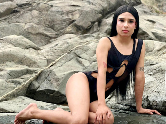 NahimaraSexy with long black hair, wearing a black cut-out swimsuit, sitting on rocky shore, showing tattoos on her right arm, and a small tattoo on her left thigh.