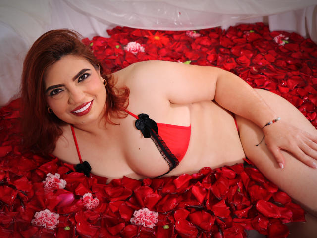 SophiaMadison with red petals, lying on her side, wearing a red bikini with black lace, smiling, showing her big tits, and red lipstick, with brown hair and a bracelet.