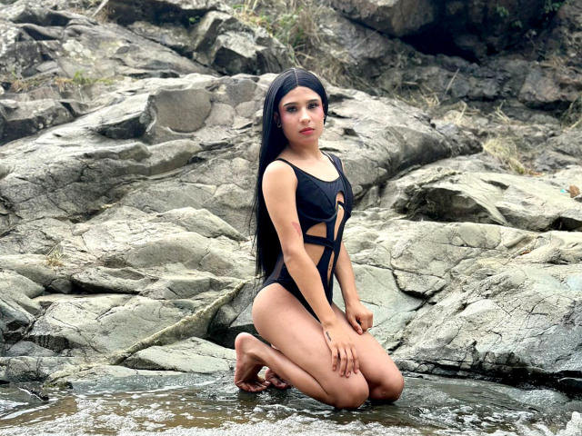 NahimaraSexy with long black hair, wearing a black cut-out swimsuit, kneeling in shallow water on rocky terrain, looking at the camera with a serious expression.
