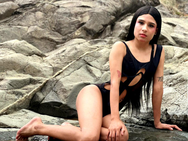 NahimaraSexy with long black hair, wearing a black cut-out one-piece, sitting on rocky ground, showing tattoos on arms, looking at camera, legs bent, bare feet, serious expression.