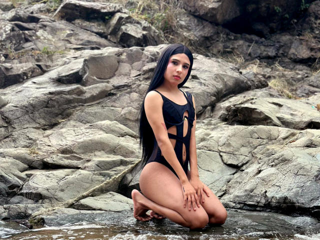 NahimaraSexy, kneeling in rocky water, wearing a black one-piece with cutouts, long black hair, and red lipstick, showing her ass and small tits, with hands on her thighs.