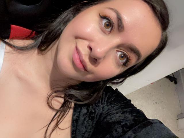 KimKolly has brown eyes, long dark hair, and a slight smile. She's wearing a black velvet top, showing her bare shoulders. Her pink lips are slightly parted.
