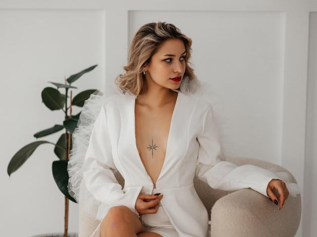 ShawtyMaria with blonde hair, wearing a deep V white blazer, exposing her tits, a star tattoo on her chest, sitting in a beige chair, red lipstick, looking to the side.