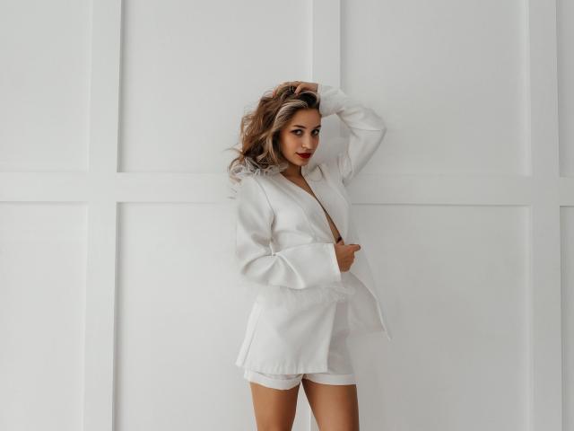 ShawtyMaria with wavy blonde hair, wearing a white, button-down, loose-fitting, short jacket, standing against a white wall, hand in hair, looking at camera with red lipstick.