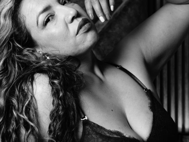 Mariehss with curly hair, dark lipstick, and lace bra, her hand near her face, showing her tits and a slightly open mouth in a black-and-white photo.