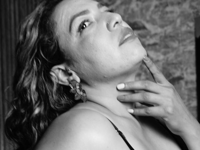 Mariehss com cabelo cacheado, piercing no nariz e brincos de borboleta, inclinando a cabeça para trás, tocando o pescoço com os dedos bem cuidados, vestindo uma blusa preta com alças, em uma foto em preto e branco..