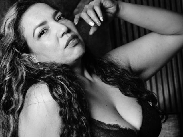 Black-and-white photo of Mariehss with wavy hair, looking down, arms raised, wearing lace bra, showing large tits, with a serious expression, hands near face, nails painted.