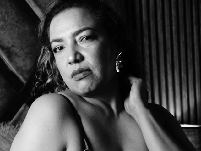 Photo en noir et blanc de Mariehss, avec des cheveux ondulés, une expression sérieuse, une main sur le cou, portant un haut sans manches, des boucles d'oreilles en forme de papillon, avec des seins visibles, et une expression légèrement boudeuse..