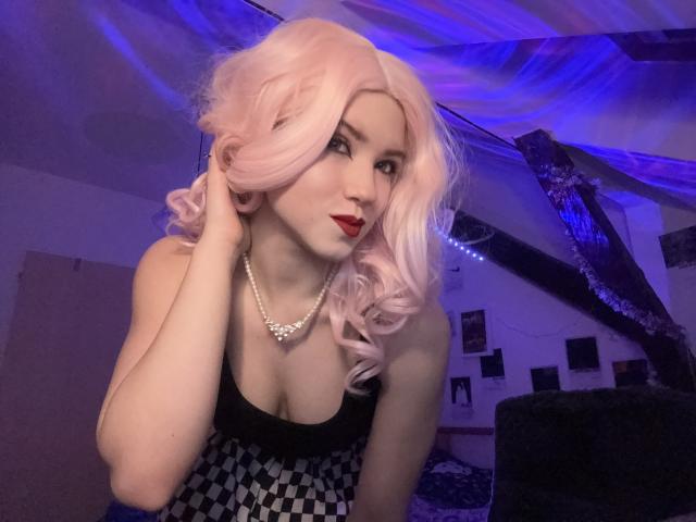 EvaxFoxy with pink curly hair, red lipstick, black and checkered dress, pearl necklace, touching hair, showing cleavage, in dimly lit room with purple and blue lights.