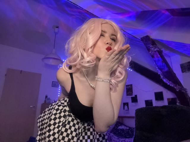 EvaxFoxy with pink curly hair, black tank top, checkered skirt, red lipstick, and white gloves, kissing her hand, showing her tits, in a room with purple lights.