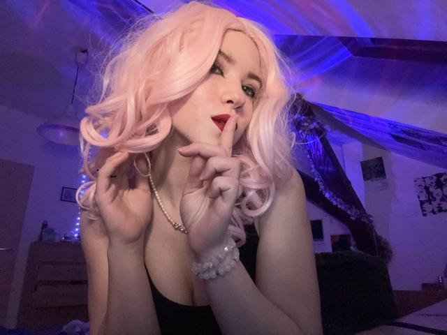 EvaxFoxy with pink curly hair, black dress, and pearl necklace, holding fingers to her red-lipped mouth, showing off her tits. Blue and purple lights in the background.