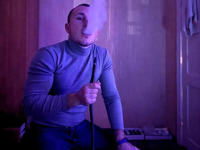 FortuneNathan, bald, wearing a gray turtleneck, holds a smoking hookah with a large, white, swirling puff of smoke covering half his face. He sits in purple light.