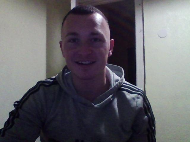 FortuneNathan, short-haired dude, smiling, wearing a gray hoodie with black stripes, white earbuds in, dimly lit, door in background, slightly blurred, casual, relaxed, short haircut.