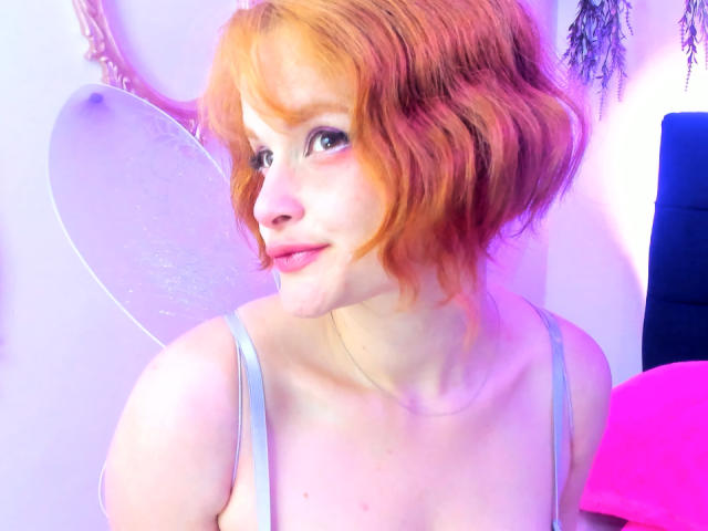 DanaStone has orange-red, wavy, short hair, light skin, and wears a gray tank top, with small tits and a slight smile, fairy wings in the background.