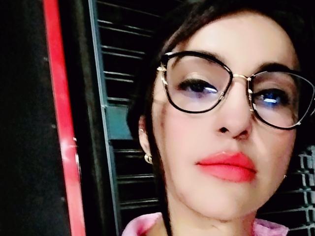 LuluFrancaise with red lips, glasses, and dark hair, showing a serious face, has a small nose and is wearing a pink shirt. Her tits aren't visible.