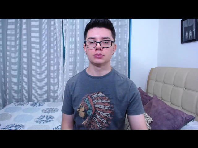 JacobWinds, short Asian dude with black glasses and a topknot, wearing a gray t-shirt with a Native American head graphic, standing in a bedroom with a beige headboard.