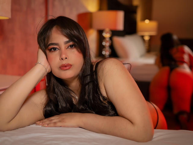 BrianaRogers, with long black hair and red lipstick, leans on a bed, her head resting on her hand, showing her tits in a black top, looking sexy.