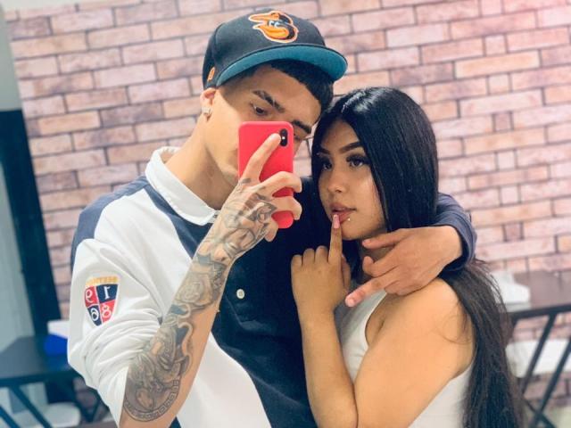 David und Laura mit Tattoos, David trägt ein weißes und schwarzes Hemd, eine Mütze, hält ein rotes Telefon, Selfie. Laura, langes schwarzes Haar, weißes Top ohne Ärmel, Finger an den Lippen, steht nah, Arm um David gelegt..