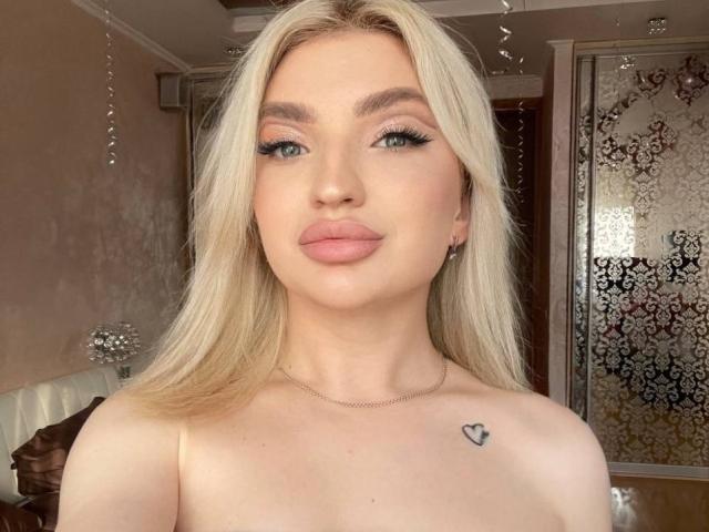 PamellaSweet with blonde hair, blue eyes, and pink lipstick, looking at the camera. She's topless, showing a small heart tattoo on her right breast.
