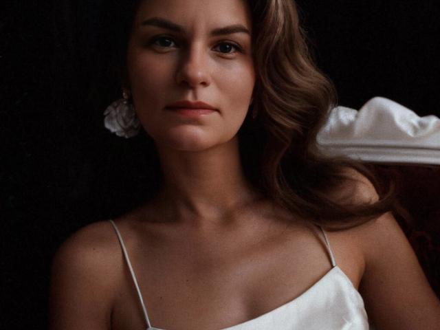 BluesStar with wavy brown hair, white spaghetti strap dress, and subtle makeup, staring intently with a serious expression, her small tits barely visible under the dress.