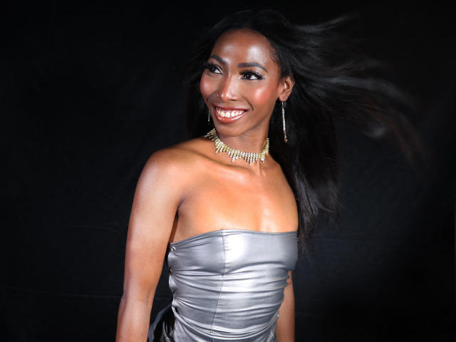 SabrinaDevil with shiny silver strapless dress, gold necklace, smiling, dark skin, long black hair blowing, wearing dangling earrings, standing against black background.