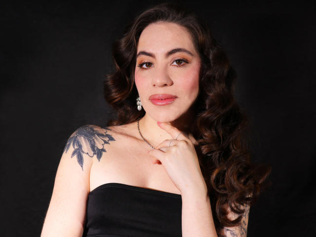 LuciaPerth with curly brown hair, black strapless top, white earrings, tattoo on left shoulder, pink lipstick, and ringed finger, against a dark background, looking at camera.