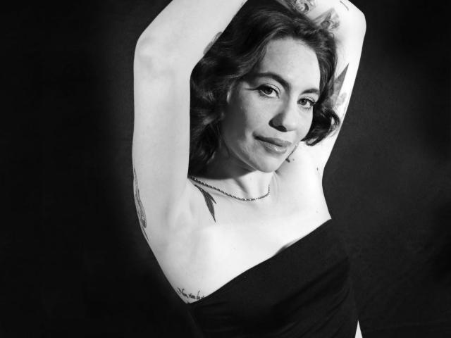 Black and white photo of LuciaPerth with arms raised, showing her bare tits and a small tattoo on her right side. She's wearing a strapless black top and a necklace.