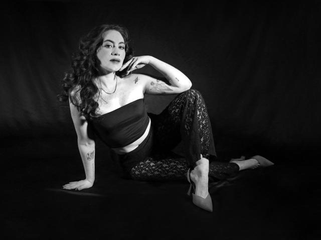 LuciaPerth with curly hair, tattooed arm, wearing a strapless top, lace pants, and pointed heels, sitting with one hand on the floor, looking confident. Black and white photo.