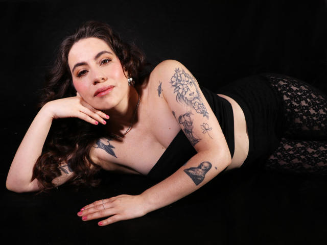 LuciaPerth with curly brown hair, lying on black background, wearing black strapless top and lace leggings, showing tattoos on arms and chest, looking at camera, pink nails.