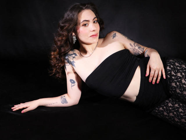 LuciaPerth with curly brown hair, black strapless top, and lace pants, showing tattoos on arms and legs, lying on black background, looking at camera with pink lipstick.
