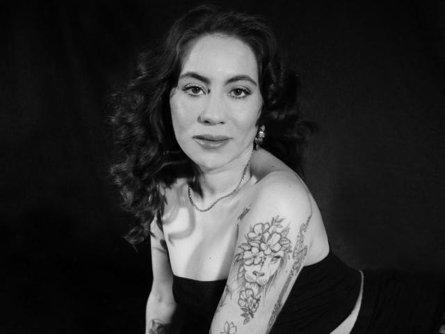 LuciaPerth with curly dark hair, wearing a strapless black top, showing a tattooed arm with flowers on her shoulder, and a necklace, looking directly at the camera. Black and white.