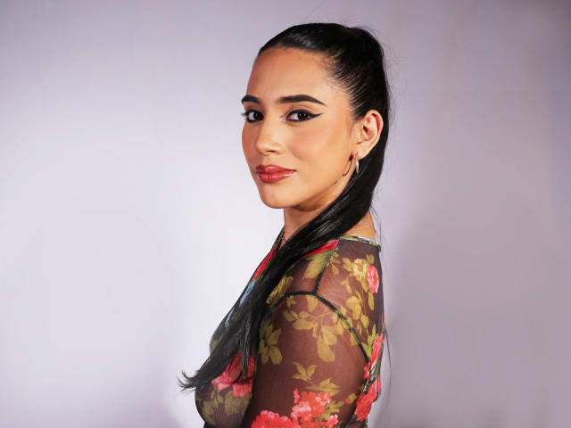 KristenHotest with long black ponytail, wearing sheer floral top, showing her tits, red lipstick, winged eyeliner, and gold hoop earrings, smiling at camera, light skin, brown eyes.