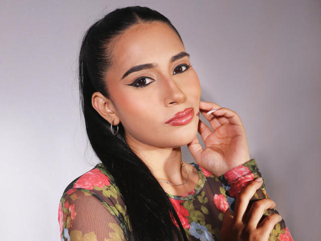 KristenHotest has dark hair in a ponytail, wearing a floral sheer top, with glossy pink lips, winged eyeliner, and hoop earrings, touching her cheek.