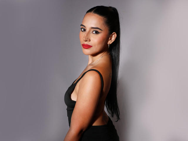KristenHotest with black hair in a ponytail, wearing a black dress, showing her back and sideboob, red lipstick, and a silver necklace, looking over her shoulder.
