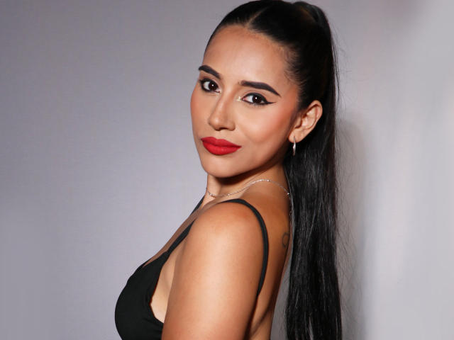 KristenHotest with dark hair in a high ponytail, red lipstick, black dress, and silver hoop earrings, looking over her shoulder, showing her bare back and small ass.