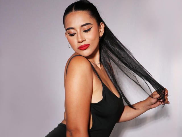 KristenHotest with long black hair, red lipstick, and black tank top, holding her hair, showing her tits, and looking down with eyeliner. She's cute and sexy.