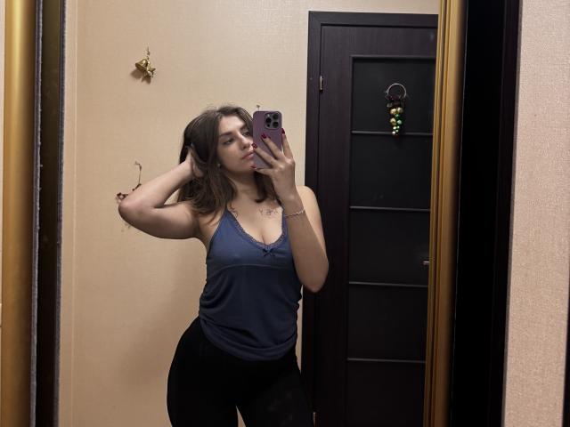 Kametia with medium tits, wearing a blue tank top and black pants, taking a mirror selfie, hand in hair, beige wall, dark door, and gold frame in the background.