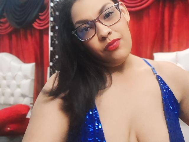 HazanFoxy with glasses, blue sequined dress, showing tits, red lips, medium skin, black hair, slightly overweight, confident, bedroom setting, red curtains in background.
