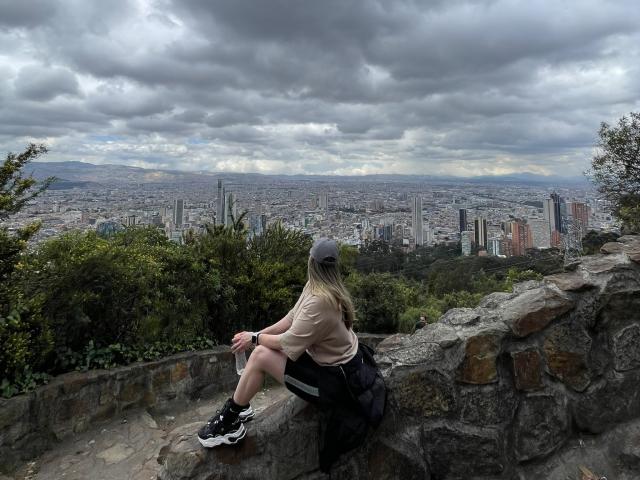 JennyLawry, con un trasero grande, sentada en un muro de piedra, vistiendo una camisa beige, pantalones cortos negros, una gorra gris, una mochila negra y zapatillas negras, contemplando la ciudad desde un punto de vista elevado, rocoso y verde..