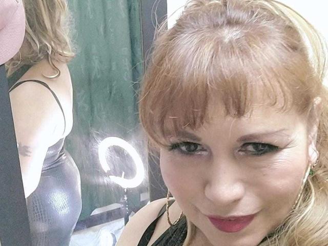 Kazsandra with blonde hair and bangs, wearing a black shiny dress, showing her ass in a mirror selfie, smiling with red lipstick, ring light in background.