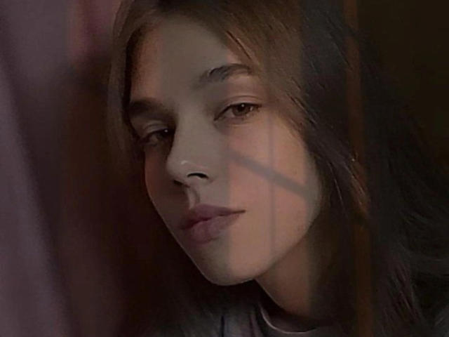 MelissaCrayzy has a smug expression, long brown hair, and a shadow across her face in a dimly lit image. Her eyes are half-closed, and she's wearing a dark shirt.