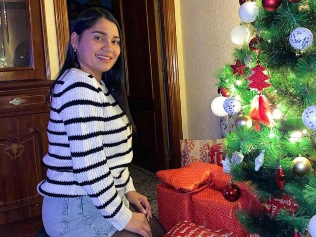 NicollCooper with long black hair, striped white and black sweater, and gray skirt, smiling next to a Christmas tree with red and white ornaments, and red gifts.