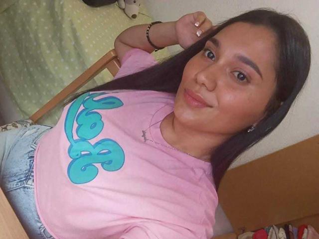 NicollCooper has dark long hair, a cute smile, and is wearing a pink shirt with blue letters, showing her small tits and wearing denim shorts.