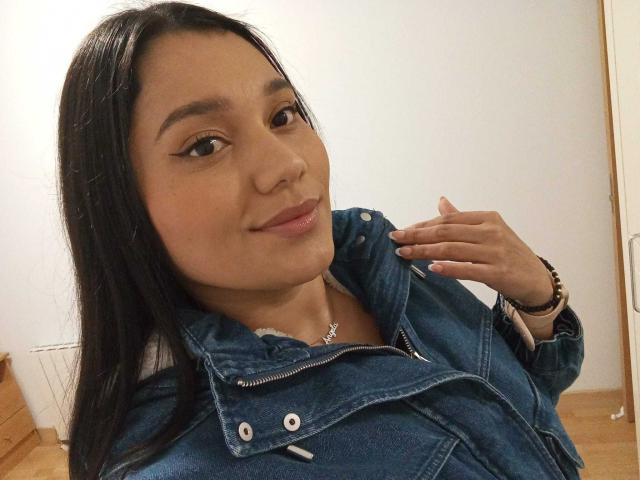 NicollCooper with long black hair, wearing a denim jacket, smiling slightly, hand near face, brown eyes, light skin, subtle pink lipstick, and a necklace, in a casual selfie.
