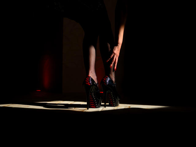 KamiLove's legs, wearing black studded heels with red laces, stand in shadow with sunlight highlighting her pale skin and red-painted nails, bending slightly at the knees.