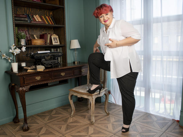 GrannyEva met rood krullend haar, een witte blouse, zwarte broek met strepen en zwarte hakken, staat met één been op een stoel, waarbij haar achterkant zichtbaar is, in een kamer in vintage-stijl..