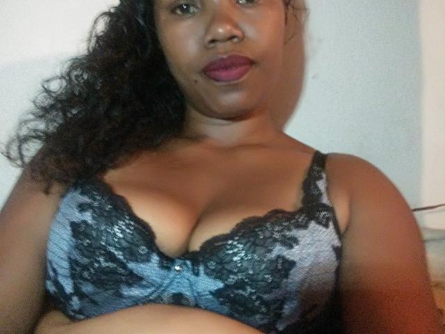 Tiane, com cabelo preto encaracolado, batom roxo escuro, vestindo um sutiã de renda azul e preto, mostrando seus seios grandes e redondos. Ela tem um tom de pele marrom médio e um olhar sério..