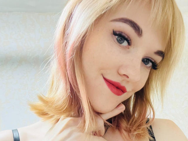 AnnaMalins69 with blonde hair, red lipstick, and black eyeliner, smiling at the camera, hand near chin, blue eyes, and a cute, sexy look, showing off her pretty face.
