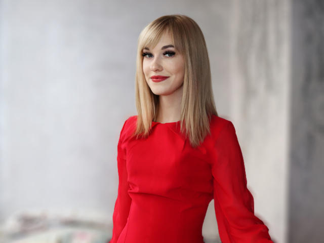AnnaMalins69 with blonde hair, red dress, and subtle smile, standing with her tits slightly visible, wearing red lipstick, in a softly lit room, looking cute.
