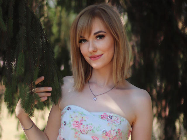 AnnaMalins69 with blonde bob, floral strapless top, holding green pine branch, smiling, pink lipstick, necklace, subtle makeup, sunlight, soft shadows, upper body, cute, sexy, natural light.